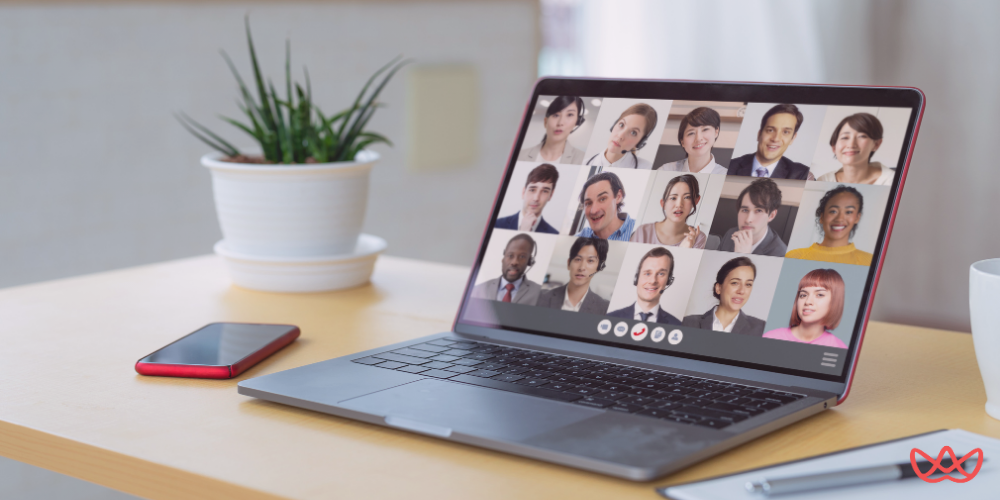 A wide shot of a modern laptop on a wooden desk, displaying a grid of diverse faces in a video conference call, with a green plant and a smartphone beside it.