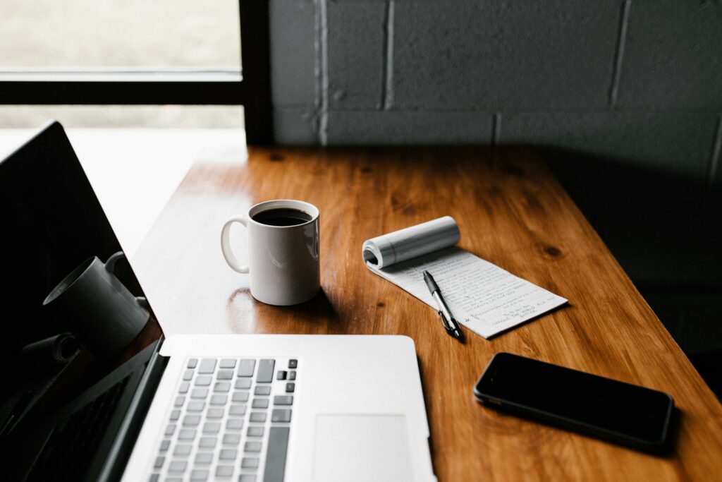 A laptop, a white coffee mug, a rolled-up notebook with a pen, and a smartphone on a wooden desk next to a window with a brick wall.