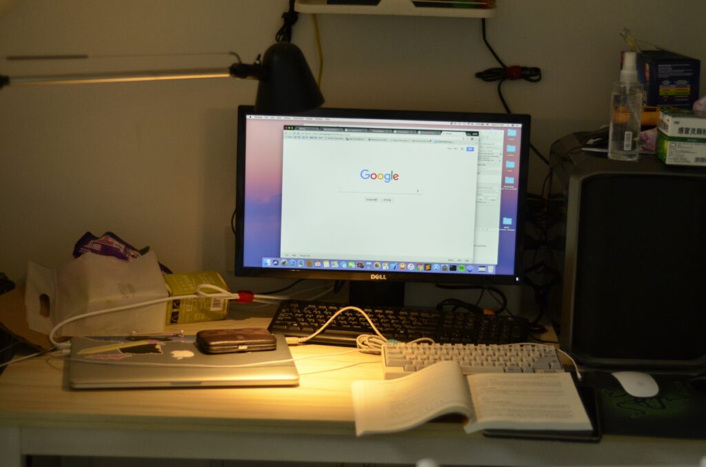A cluttered desk with a large computer monitor displaying the Google search page, a laptop, multiple keyboards, an open book, and various electronic accessories.