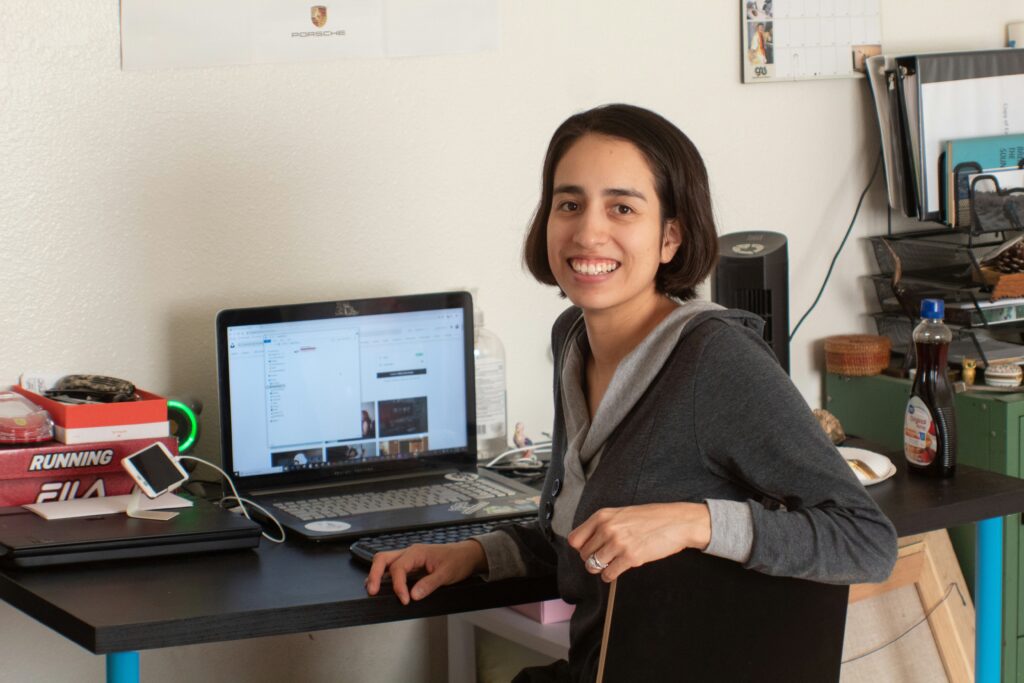 A woman with dark hair, wearing a gray hooded top, smiling and looking directly at the camera while seated at a desk with a laptop and various office items in the background.