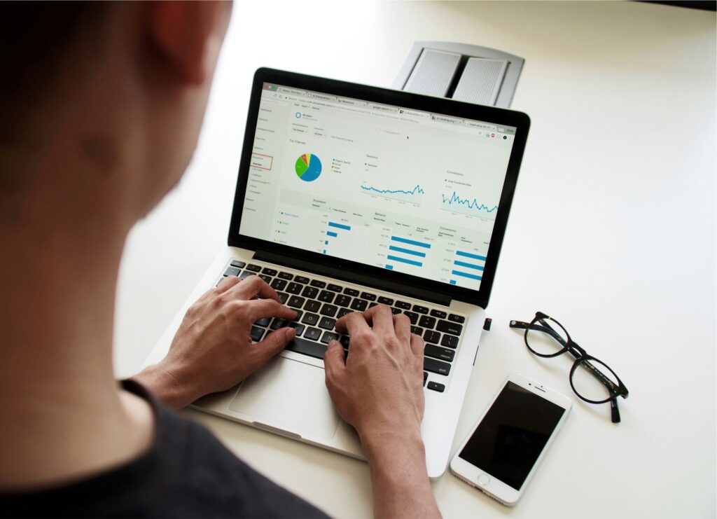 Overhead shot of a person's hands on a laptop keyboard and touchpad, with a smartphone, notebook, and stack of books on a desk, suggesting a workspace for managing finances or business operations. The laptop screen shows the text "Top Outsourced Accounting Services & Trends in 2025.