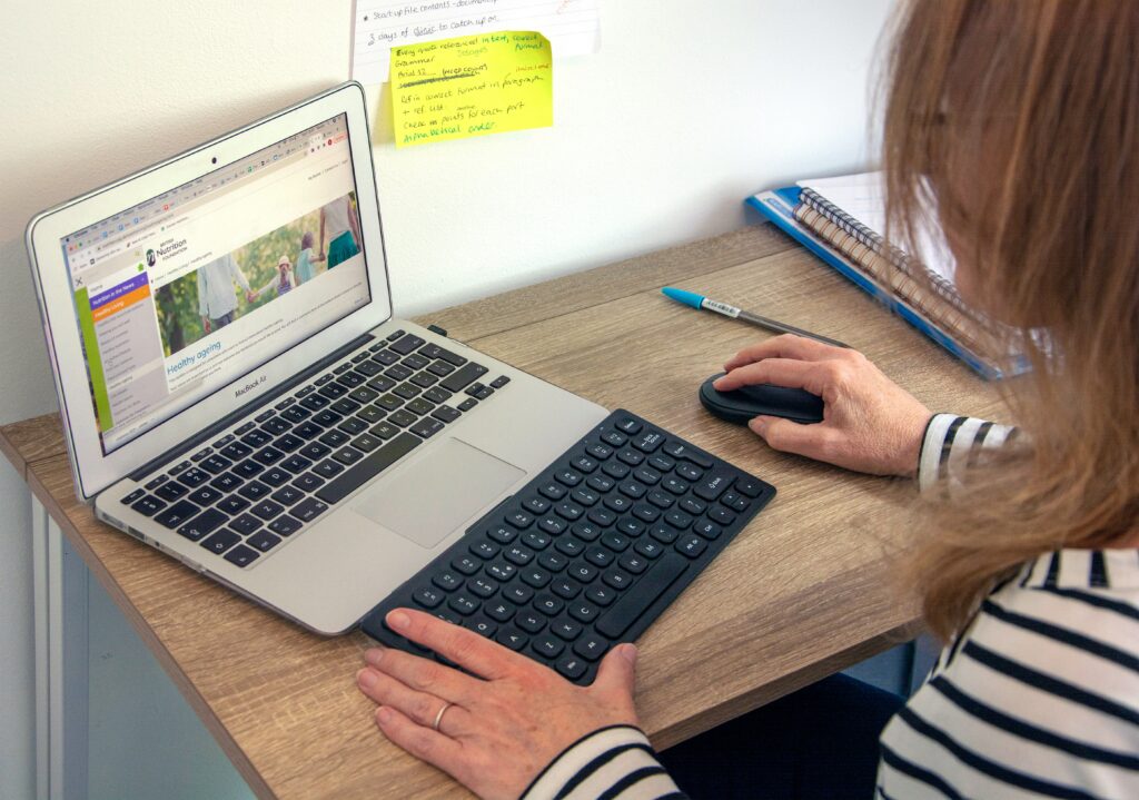 A person from behind, wearing a red beanie and a blue jacket, typing on a laptop with the Google search page open on the screen, implying internet research or digital work in an outdoor setting.