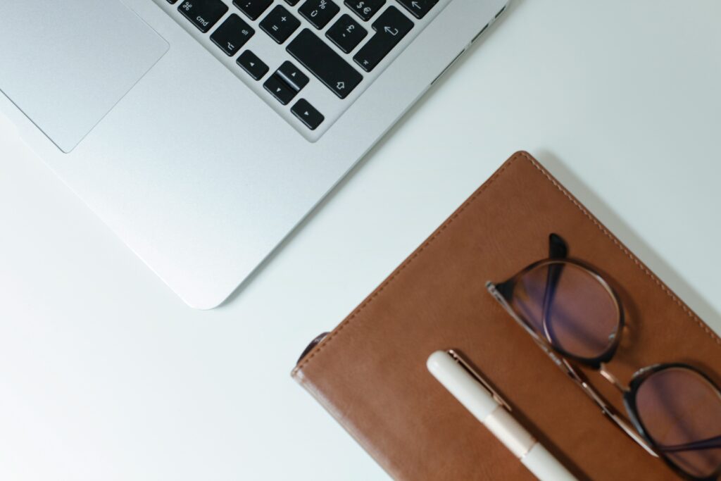 A close-up of a person writing in a brown paper bag, with a laptop, tablet, scattered financial documents, and a coffee cup on a wooden desk, suggesting a busy work environment focused on data or finances.