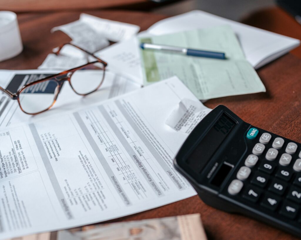 A person's hands typing on a laptop keyboard, with US dollar bills, a smartphone displaying a calculator app, and documents with charts scattered on a wooden desk, symbolizing financial management. The image also contains the text "In-House Payroll vs Outsourcing Which is Right for Your Business."