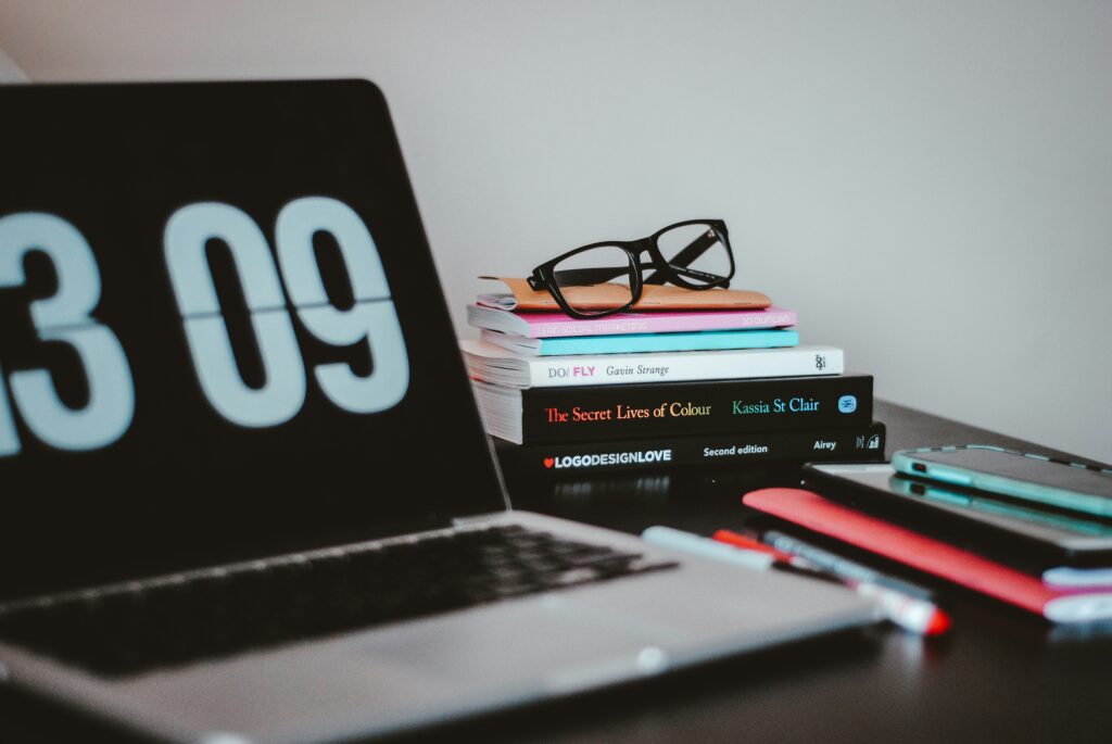 A tidy desk setup featuring a laptop with a digital clock displaying "3 09", a stack of books, eyeglasses, and a smartphone, suggesting a focused and organized workspace.