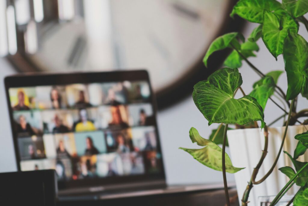 A plant in the foreground, slightly out of focus, with a laptop in the mid-ground displaying a video conference grid of diverse people. A clock is visible in the background, also out of focus.