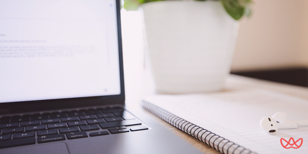 A close-up view of a laptop screen displaying text, next to a spiral-bound notebook and wireless earbuds on a desk. A potted plant is blurred in the background, suggesting a focused workspace often seen in a top product management recruitment agency.