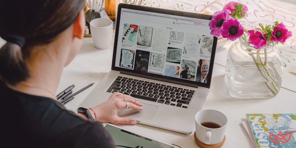 A person from behind views a laptop screen displaying a grid of creative designs, with a title card visible that reads "Top Visual Designers to Watch in 2025." A vase of pink flowers and a cup of coffee are also on the desk.