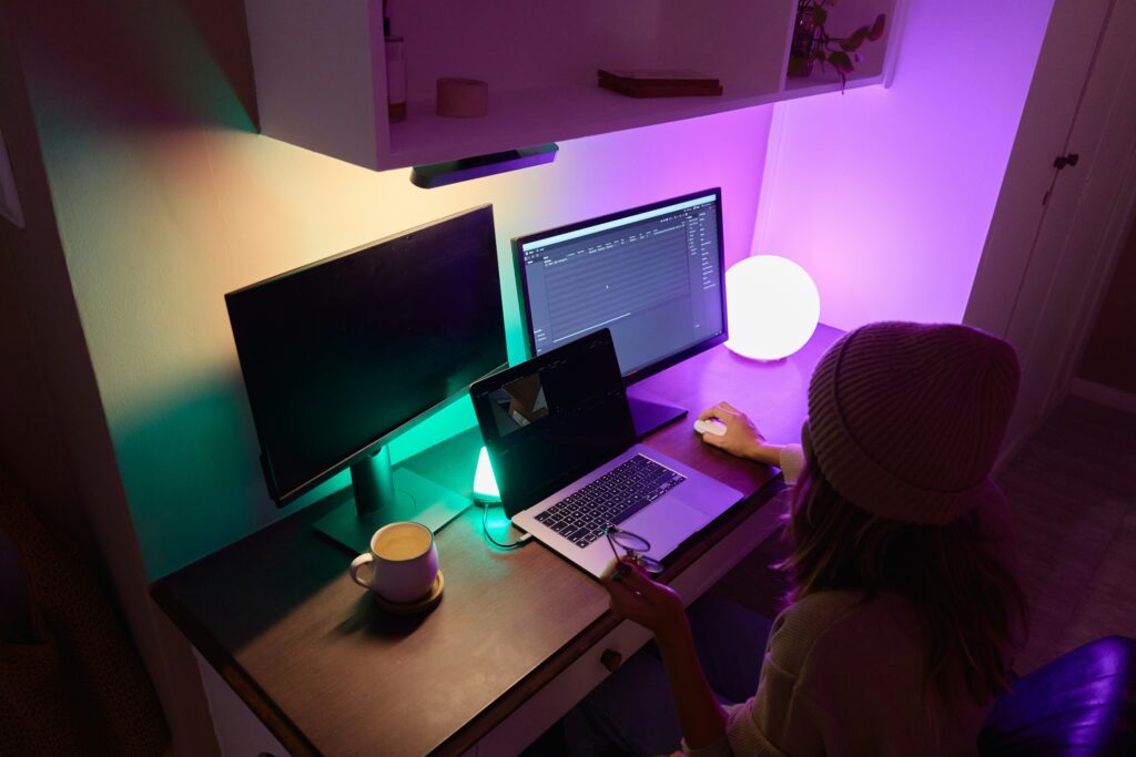 A person from behind works at a multi-monitor desk setup bathed in colorful ambient lighting, with a laptop, two larger monitors, and a cup of coffee. This vibrant and creative workspace is indicative of environments where top visual designers might work.