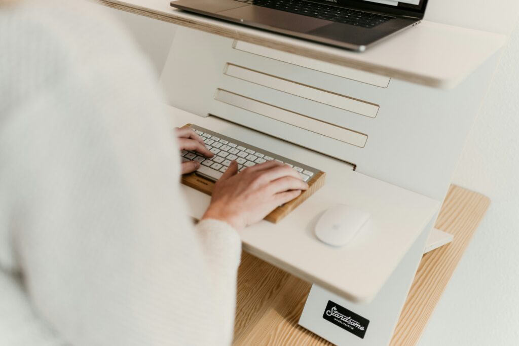 A person stands and types on a keyboard at a modern wooden standing desk converter, with a laptop visible on the upper tier and a white mouse below. This professional and organized setup is common in dynamic workplaces, including a top recruitment agency.